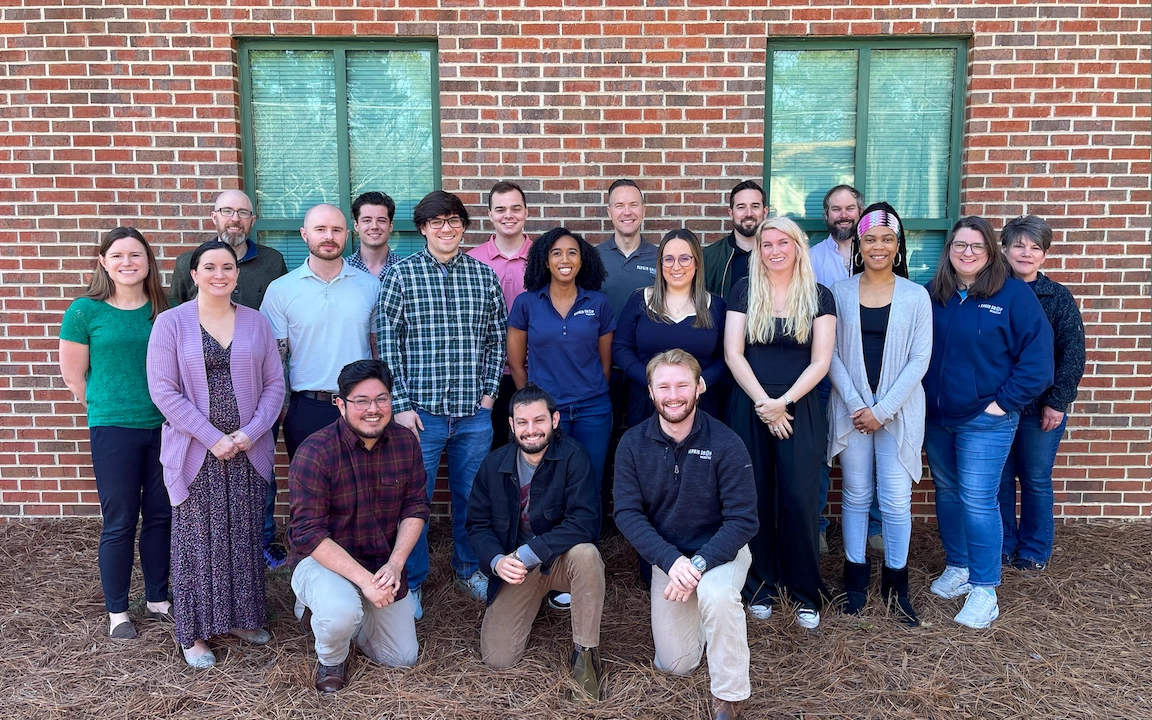The Repair Shop Websites team, based in the USA, poses for a group photo in front of a red brick building with two green-tinted windows. 